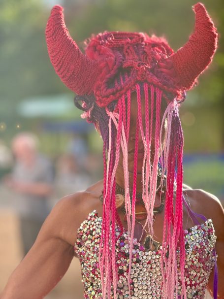Person with red horns and colorful hair, wearing a beaded outfit, facing away.
