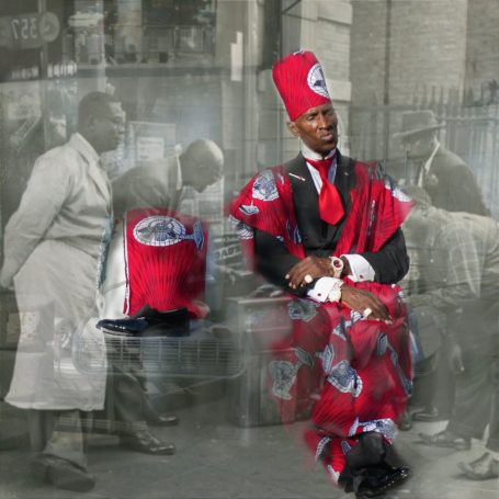 Red Ensemble A man in a red patterned outfit and hat sits confidently in a blurred vintage urban setting in the background.