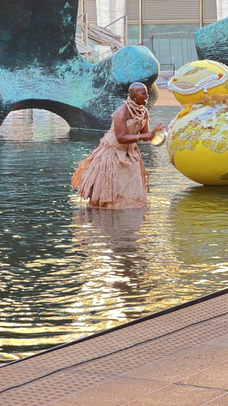 A figure in a flowing dress interacts with a large yellow sculpture in a reflective pool.