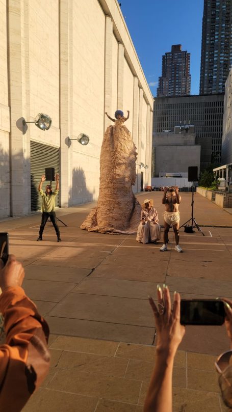 A large, textured sculpture with people gathering around it outdoors.
