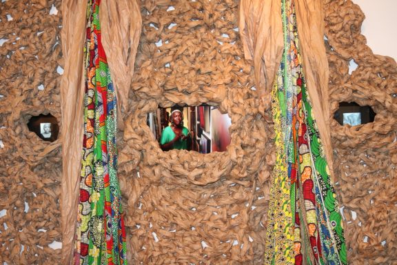 Colorful fabrics and objects are woven into a textured, earthy wall installation.