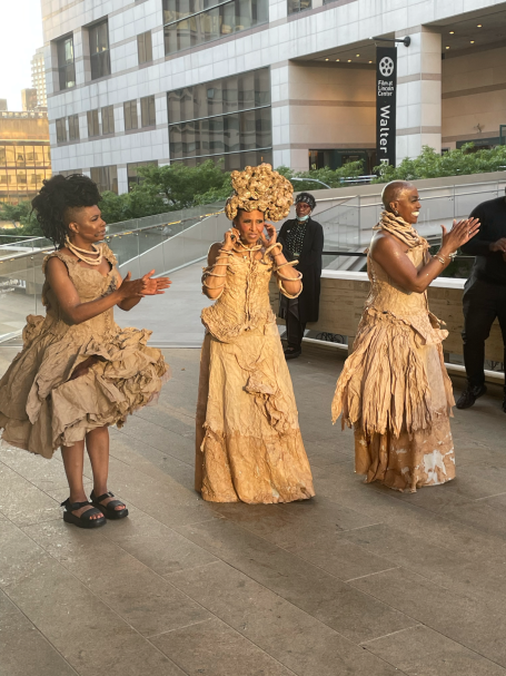 Three women in elaborate, textured dresses made of light materials, clapping together outdoors.