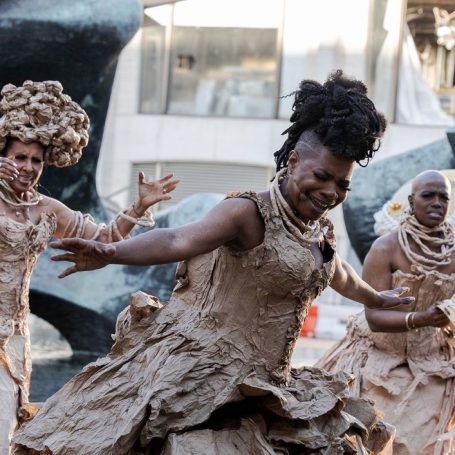 Three dancers in textured, brown costumes perform joyfully outdoors.