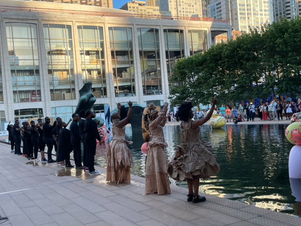 People in formal attire and elegant dresses gather by a reflecting pool at a public event.
