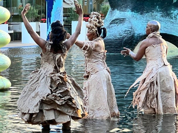 Three dancers in elegant, textured dresses perform near water, showcasing artistic movement.