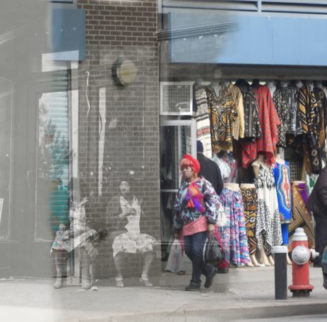 There No More A busy street scene with colorful clothing displayed and a woman walking by.