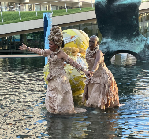 Two performers in elaborate costumes dance in water near a large, colorful sculpture.