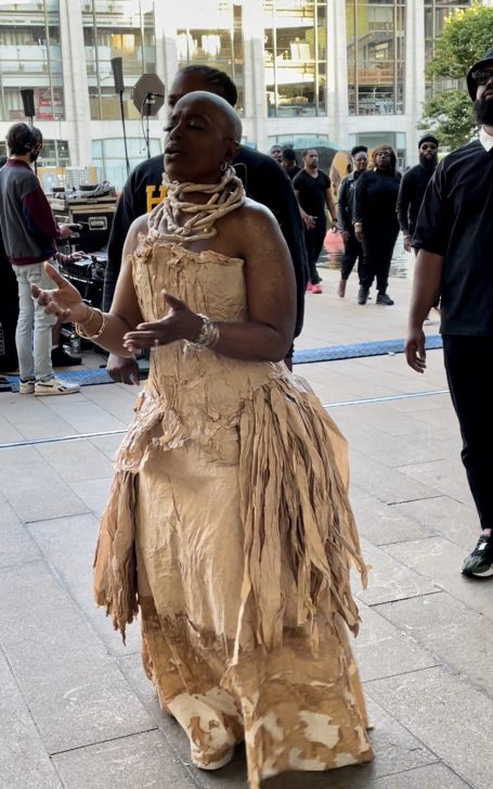 A person wearing a beige, textured dress and accessories, standing among a crowd.