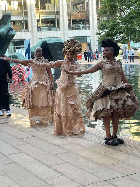 Three dancers wearing elaborate, textured costumes pose gracefully near a reflective surface.