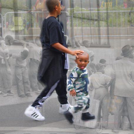 Lil Man Two children walking together, the taller one has a hand, on the smaller child's head while with blurred figures in the background.